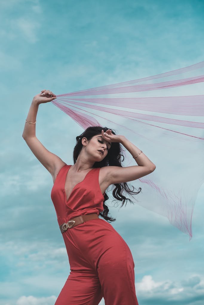 A woman poses outdoors in a striking red jumpsuit with sheer fabric against a vibrant sky.