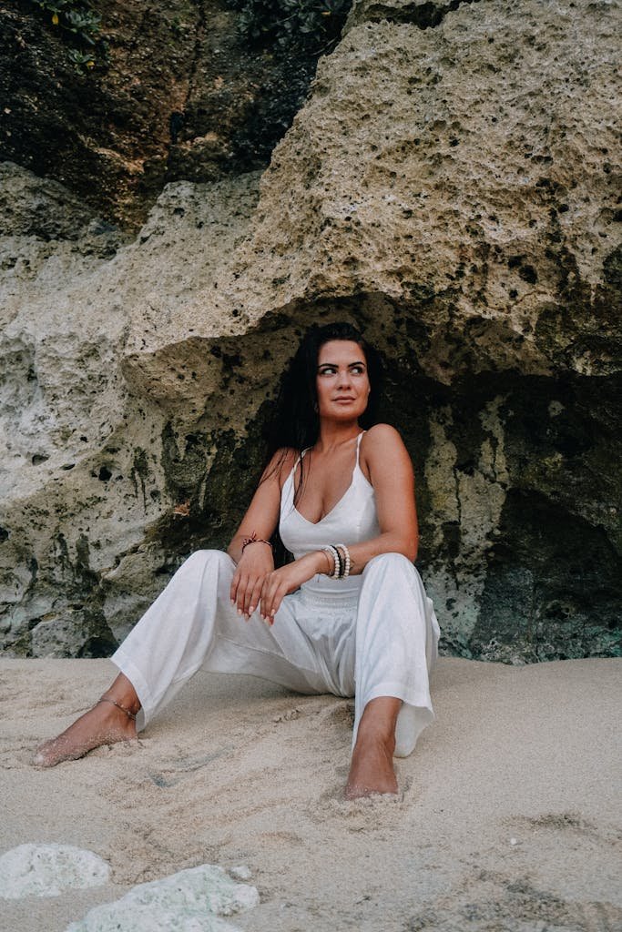 Woman in white jumpsuit sitting on sandy beach under rock formation, serene and contemplative.